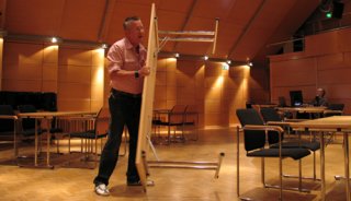 Seppo kantaa pöytiä kamarimusiikkisaliin. Taustalla Heikki Tuuli lataa koneelle uusimmat kuvat kamerasta. Kuva: Kaisa Erola/YLE.