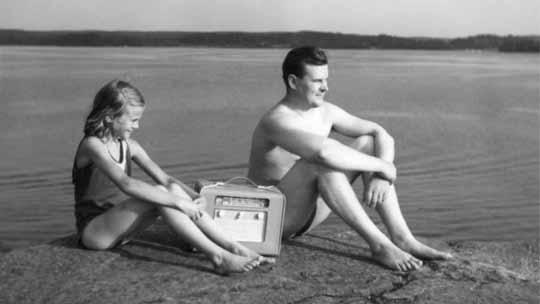 Isä ja tytär kuuntelevat matkaradiota veden äärellä. Kuva sarjasta Kun radio tuli meille. Kuva: YLE Kuvapalvelu, 1950-luku. Isä ja tytär kuuntelevat matkaradiota veden äärellä. Kuva sarjasta Kun radio tul